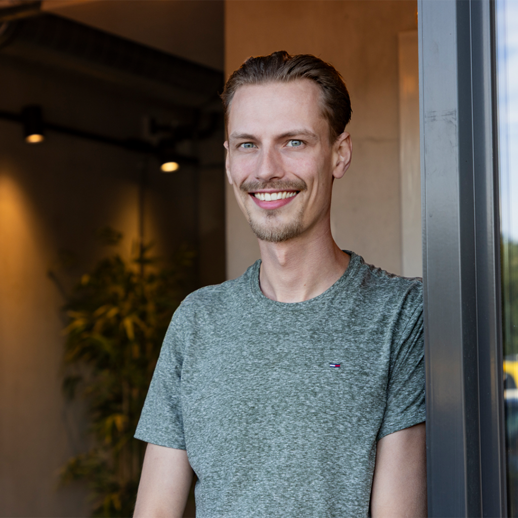 Glimlachende man in grijs shirt bij deur.